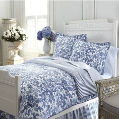 a bed with blue and white comforters in a room next to a chair, table and flowers