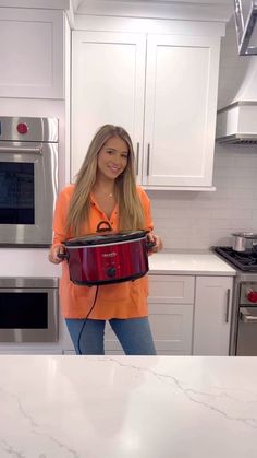 a woman in an orange shirt is holding a red crock pot on the kitchen counter