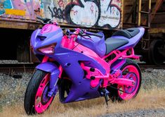 a purple and pink motorcycle parked in front of a train track with graffiti on it