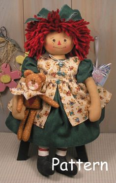 a doll with red hair holding a teddy bear and wearing a green dress is sitting on a chair