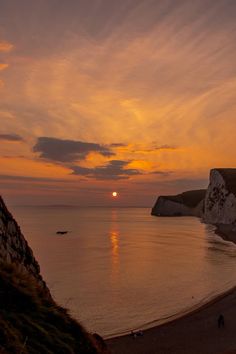 the sun is setting over the ocean with people walking on the beach and cliffs in the background