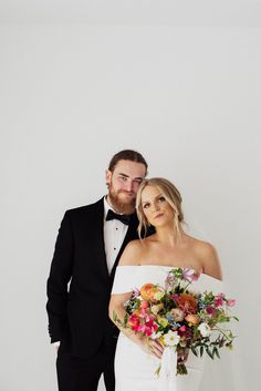 a bride and groom pose for a wedding photo in front of a white wall wearing tuxedos