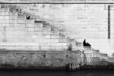 black and white photograph of two people sitting on steps next to the water in front of a stone wall