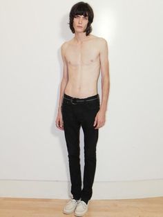 a shirtless man standing in front of a white wall wearing black pants and sneakers