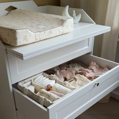 an open drawer in a white dresser with baby clothes inside it and a crib on the other side