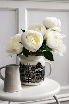 white flowers in a vase sitting on a table next to a gray mug with handles