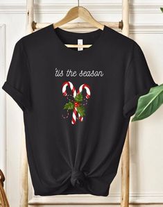 🎄Celebrate the holidays in style with this Tis The Season T-Shirt! Featuring two classic candy canes crossed together with a sprig of holly and berries, accompanied by the festive phrase 'Tis the Season in elegant white script font, this shirt is the perfect blend of sophistication and holiday cheer. 🍬✨ Ideal for Christmas parties, family gatherings, or cozy holiday shopping trips, this stylish t-shirt adds a touch of festive charm to your wardrobe. The design captures the essence of the seaso New Year Holiday Crew Neck T-shirt, Casual Black T-shirt For Festive Occasions, Black Casual T-shirt For Festive Occasions, Black Holiday Tops With Letter Print, Black Holiday Top With Letter Print, Black Tops With Letter Print For Holiday, Casual Black Tops For New Year, Festive Black T-shirt With Letter Print, Black Top As Christmas Gift