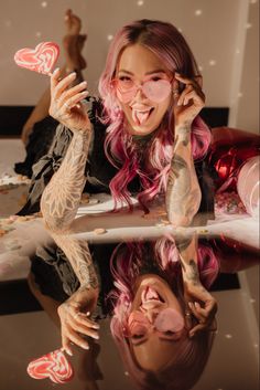 a woman with pink hair and tattoos is reflected in a mirror while she holds up her heart shaped lollipops