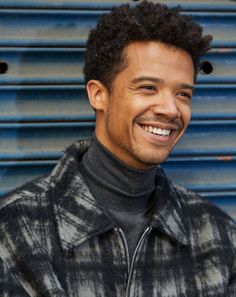 a smiling man in front of a blue wall wearing a black and white jacket with his hair pulled back