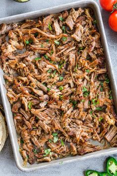 shredded pork in a baking pan with tomatoes and green peppers on the side, next to pita bread