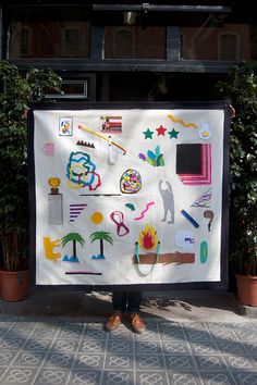 a person standing next to a large poster with various items on it and plants in the background