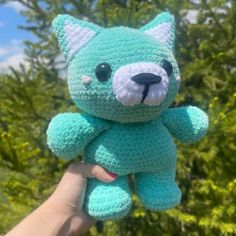 a hand holding a small blue crocheted teddy bear in front of some trees