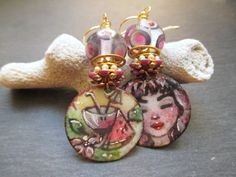 These earrings feature enameled copper charms by TheDreamCatchersCo in Bulgaria. The round charms are illustrated with a pink watermelon martini flanked by tropical fruits on one charm, and young woman with pink hair wearing watermelon earrings on the other. Above the charms are purple rhinestone and enamel flower bead caps followed by pearl spacers set in gold.  Next are open work gold saucer beads. These support stunning lamp-work glass beads by Unventdeliberte in France. The lamp-work beads are are clear with purple spots rimmed in white and grey. Bi-cone brass spacers finish off these darling cocktail dangles, which hang from simple gold ear wires and measure 2 inches long. These earrings will be sent to you attractively packaged in a gift box with ribbon. You can check out more of my Whimsical Enamel Jewelry For Jewelry Making, Whimsical Enamel Earrings Nickel Free, Fun Nickel-free Enamel Jewelry, Nickel-free Enamel Jewelry With Fun Style, Nickel-free Enamel Fun Jewelry, Artsy Enamel Earrings As Gift, Whimsical Hand Painted Earrings, Fun Enamel Earrings For Gifts, Soldered Enamel Drop Earrings