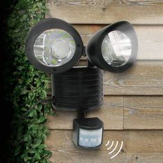 two lights on the side of a wooden building next to a bush and fence with green leaves