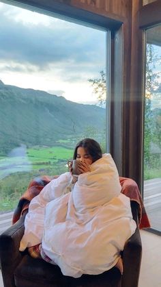 a woman wrapped in a blanket sitting on a chair looking out the window at mountains