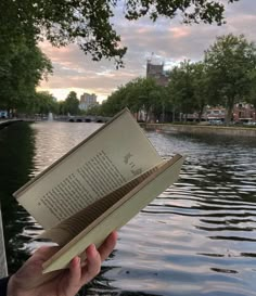 a person holding up an open book in front of water with trees and buildings on the other side