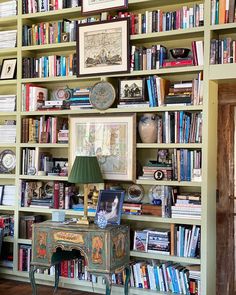 a bookshelf filled with lots of books next to a table and lamp on top of a hard wood floor