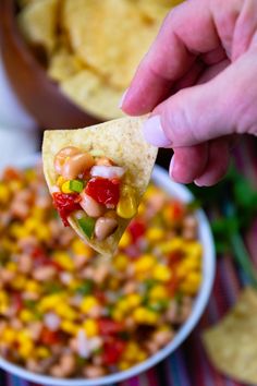 a hand holding a tortilla chip over a bowl of beans and corn salsa