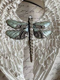 a dragonfly brooch sits on top of a white shawl draped over a piece of fabric