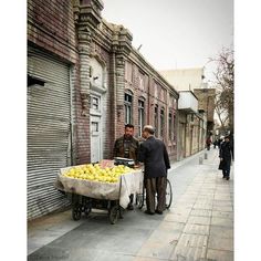 two men standing next to a wheelbarrow filled with lemons on the side of a street