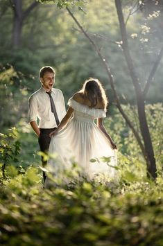 a man and woman are walking through the woods holding hands, looking at each other