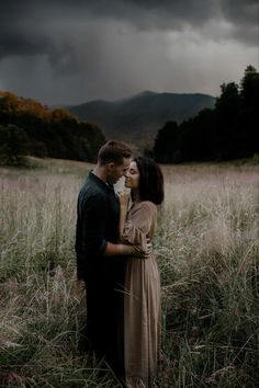 a man and woman standing in tall grass under a cloudy sky with mountains behind them