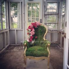 an old chair covered in moss with flowers on the seat and windows to the side