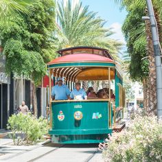 a green trolley car with people riding on it's side and palm trees in the background