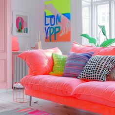 a bright pink couch with colorful pillows on it in front of a painting and window