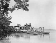 black and white photograph of people on a boat in the water near a dock with trees