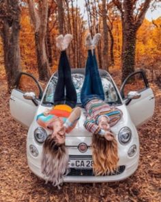 two women laying on the hood of a car in front of trees with autumn leaves