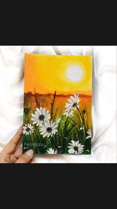 a hand holding up a small card with daisies in the foreground and an orange sky behind it