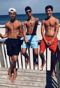 three shirtless men standing on a boardwalk at the beach with one pointing to the camera