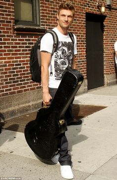 a man holding a guitar case while walking down the street in front of a brick building