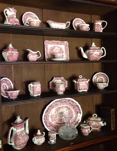 an assortment of pink and white china on display in a cabinet with wooden shelves behind it