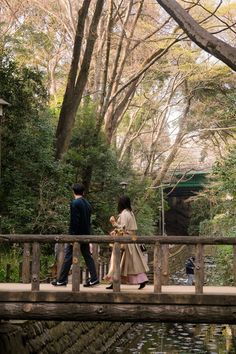 two people walking across a wooden bridge over a small river in a park with lots of trees