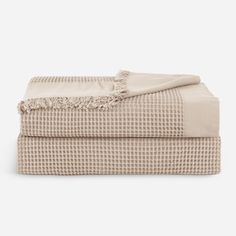 two blankets folded on top of each other, one with a fringed edge and the other in beige