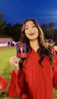 a woman in a red graduation gown holding up a card and smiling at the camera