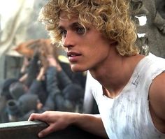 a young man with curly hair standing next to a wall and looking off into the distance