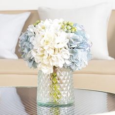 a glass vase filled with white and blue flowers on top of a table next to a couch