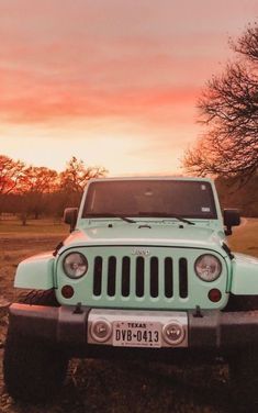 a green jeep parked on top of a dirt field next to a tree at sunset