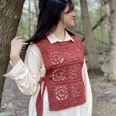 a woman standing next to a tree wearing a red crocheted vest and white shirt