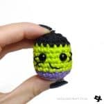 a hand holding a small crocheted green and purple ball with eyes on it