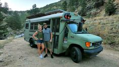 two people standing in front of a green bus on the side of a dirt road