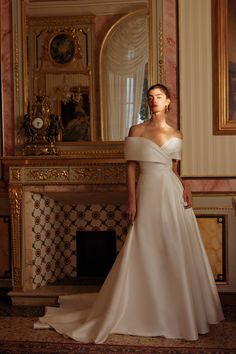 a woman standing in front of a fireplace wearing a wedding dress with an off the shoulder neckline