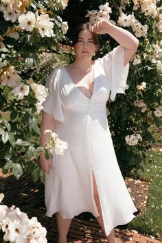 a woman in a white dress posing for the camera with flowers around her neck and hands behind her head