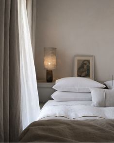 a bed with white sheets and pillows next to a lamp on a table in front of a window