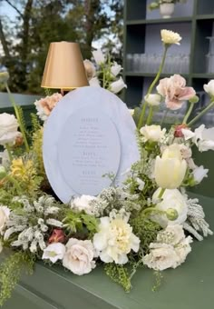 an arrangement of flowers and greenery with a plaque in the center on a table