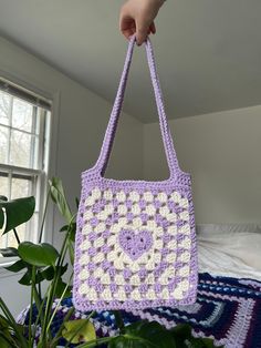 a hand holding a crocheted purple and white purse next to a potted plant