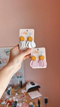 a person holding three pairs of earrings in front of a desk with other items on it